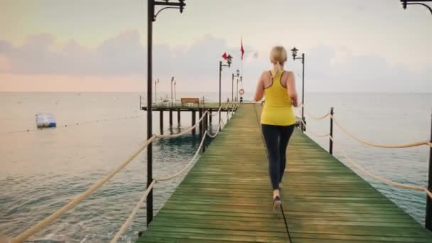 Aktive Lebensweise: Ein junges Mädchen in Sportbekleidung läuft morgens auf der Seebrücke zum Sonnenaufgang — Stockvideo