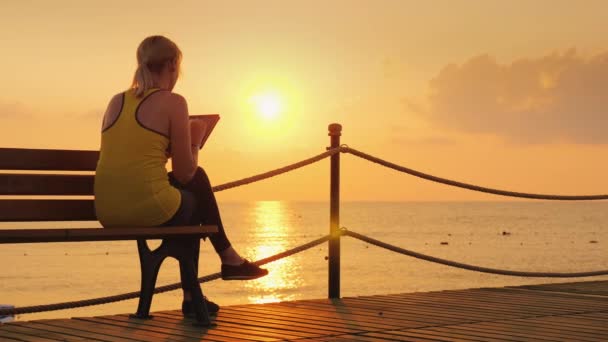 Mujer fitness disfruta de una tableta. Se sienta en un banco en un muelle contra el telón de fondo del sol naciente. Vista trasera — Vídeos de Stock