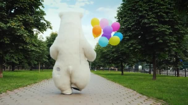 Rückansicht: Ein großer Eisbär läuft die Straße hinunter, trägt Luftballons und eine Schachtel mit einem Geschenk. Geburtstagsgeschenke und cooles Party-Konzept — Stockvideo