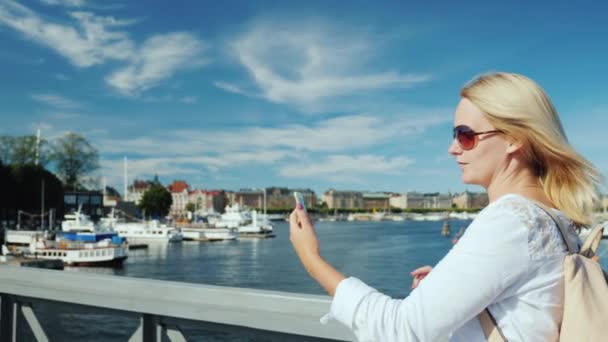 Blogger žena komunikuje prostřednictvím video chatu, chodí na most přes řeku proti pozadí řádku město Stockholm, Švédsko. Cestování blog koncept — Stock video