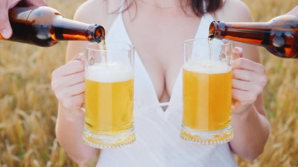 Uma mulher atraente com um belo peito segura dois copos de cerveja, em ambos os lados ela é derramada uma bebida fresca. No fundo um campo de trigo maduro. Conceito de sonho dos homens — Vídeo de Stock