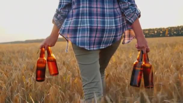 Una mujer lleva cuatro botellas de cerveza fría, camina por el campo de trigo. concepto de fantasía para hombre — Vídeo de stock