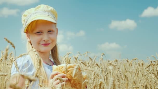 Una chica con una hogaza de pan se para en un campo de trigo, mira a la cámara — Vídeos de Stock