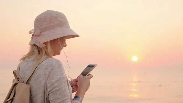 Not a minute without a phone, the girl is standing on the beach with a pink sky and sky, looking at the phone screen and listening to music on headphones — Stock Photo, Image