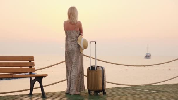 Een vrouw met een reistas en hoed in haar hand staat op de pier, de verte op zoek naar het schip. Zonsopgang boven de zee, mooie romantische video — Stockvideo