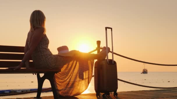 Une femme romantique en robe légère avec un sac burly s'assoit sur le quai, regarde le lever du soleil sur la mer et un navire solitaire au loin. Concept de croisière et de voyage — Video