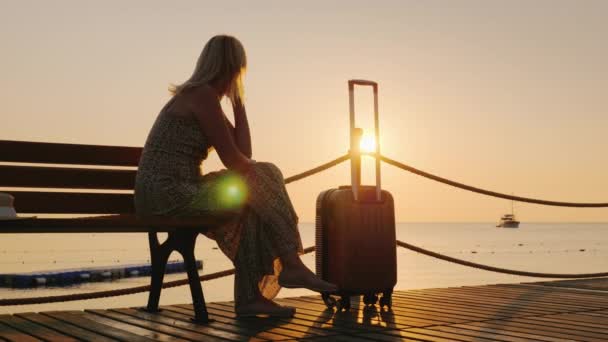 Una mujer con una bolsa de viaje se sienta en un muelle de madera, mirando hacia el amanecer sobre el mar y un barco en la distancia. Sueño con viajar y concepto de libertad — Vídeo de stock