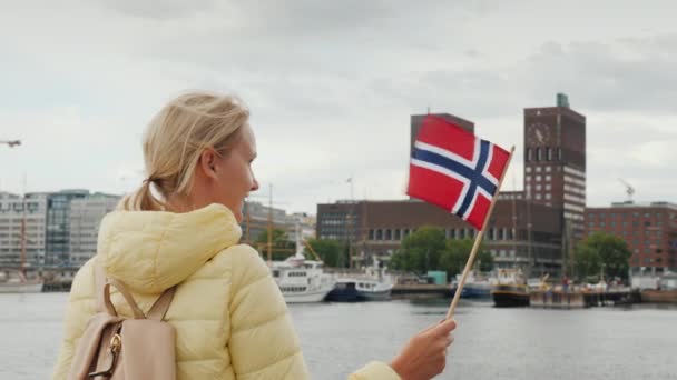 Uma turista feminina com uma bandeira da Noruega está de pé no cais, olhando para a frente para a cidade de Oslo, a capital da Noruega. Uma viagem através da Escandinávia Conceito — Vídeo de Stock