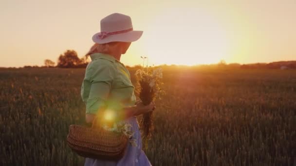 Yazlık giysiler ve bir şapka genç bir kadında günbatımında alanına yürüyor. Kır çiçekleri Sepeti taşır — Stok video