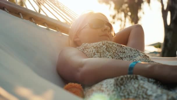 De toerist van een vrouw in een zomerjurk ontspant in een hangmat. De zon op de skyline maakt prachtige hoogtepunten in het frame — Stockvideo