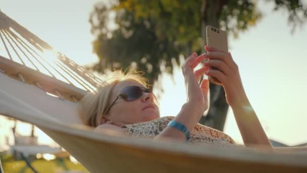 Bir kadın bir smartphone tatile kullanır, sabahın erken saatlerinde bir hamakta rahatlatır — Stok video