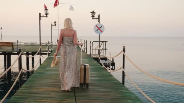 Eine Frau mit einer Tasche auf Rädern fährt über die Seebrücke in Richtung Meer, die aufgehende Sonne erhellt ihr Kleid wunderschön. auf Kreuzfahrt gehen — Stockvideo