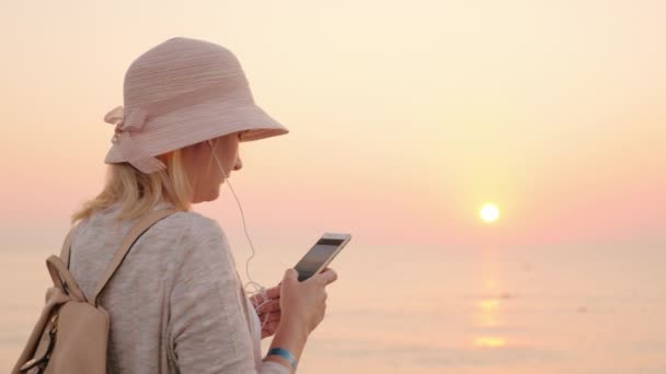 전화 없이 안 분, 여자 전화 화면을 보고 하 고 헤드폰에서 음악을 듣고 핑크 하늘과 하늘, 해변에 서 있다 — 비디오