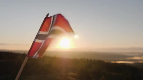 De vlag van Noorwegen fladdert in de wind, de zon schijnt prachtig door het weefsel. Op de top van de berg met een epische landschap op de achtergrond — Stockvideo