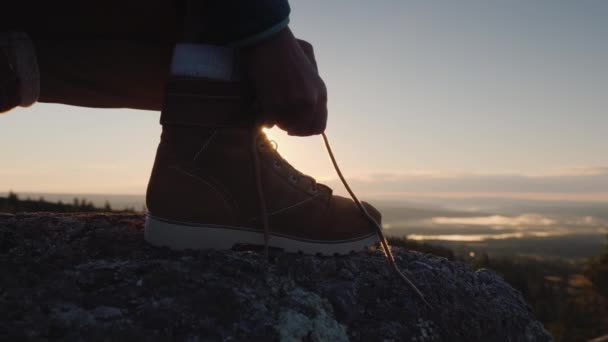 O viajante amarra os atacadores em sapatos de rastreamento. No topo da montanha contra o pano de fundo do sol nascente. O início de um grande conceito de viagem — Vídeo de Stock