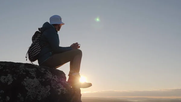 Ένας ταξιδιώτης πίνει τσάι από μια κούπα στην κορυφή του βουνού. Κάθεται σε έναν βράχο, στο παρασκήνιο ενός κόκκινου ουρανού και ουρανού. Μακριά από τον πολιτισμό, την έννοια — Φωτογραφία Αρχείου