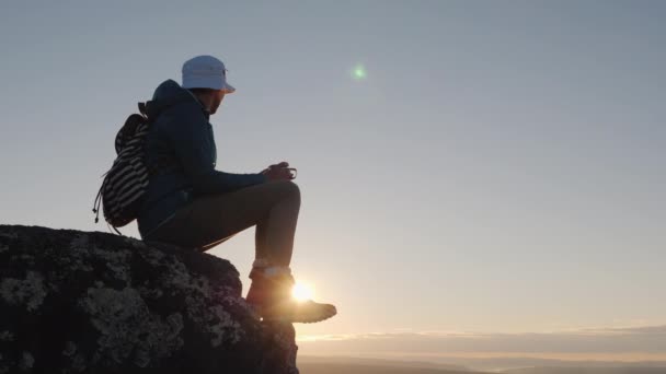 A traveler drinks tea from a mug at the peak of the mountain. Sits on a rock, in the background a red sky and sky. Away from civilization, the concept — Stock Video