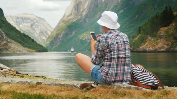 Жінка використовує смартфон на березі мальовничого фіорду в Норвегії. Завжди на зв'язку, технології на дорозі — стокове відео