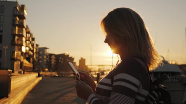 Een vrouw in hoofdtelefoon maakt gebruik van een smartphone op de pier op de achtergrond van prive-jachten. Bij zonsondergang verlicht de zon prachtig het. — Stockvideo