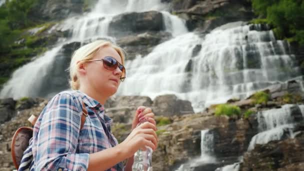 Young woman drinks water on the background of the Tvindefossen in Norway. Clean drinking water from a waterfall — Stock Video