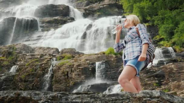 Mujer activa bebe agua limpia en el fondo de la cascada Twindefossen en Noruega — Vídeos de Stock