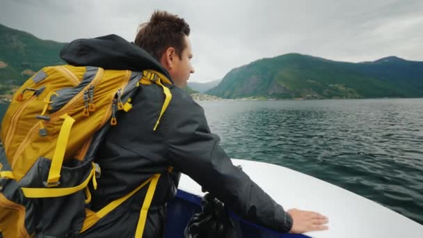 Um homem viajante fica no nariz de um pequeno navio de cruzeiro, navega em um fiorde pitoresco na Noruega — Vídeo de Stock