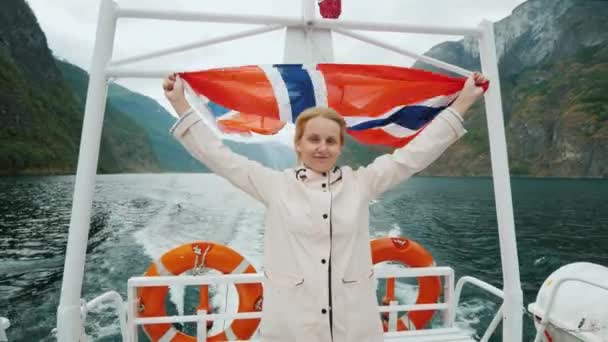Молодая женщина с флагом Норвегии стоит на корме лодки удовольствия, круиз по живописному фьорду в Норвегии — стоковое видео