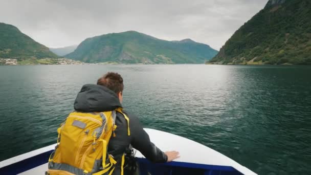 Ένας άνδρας με ένα σακίδιο που ταξιδεύει μέσα από ένα γραφικό φιόρδ της Νορβηγίας. Στέκεται στην πλώρη του πλοίου, κοιτάζοντας μπροστά — Αρχείο Βίντεο