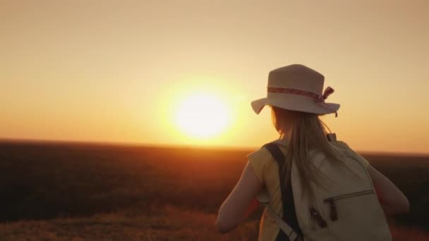 Una chica alegre corre ligeramente a lo largo de un camino rural hacia el sol poniente. Tiene un sombrero de paja y una mochila a sus espaldas. — Vídeo de stock