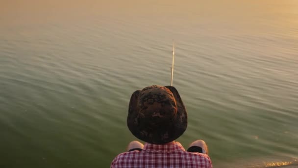 Top uitzicht: een tiener in een hoed zit op een stoel aan het meer en de vangsten van vis met een hengel — Stockvideo