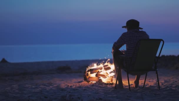 Un joven se está calentando las manos por el fuego a última hora de la noche. Se sienta junto al lago al atardecer — Vídeo de stock