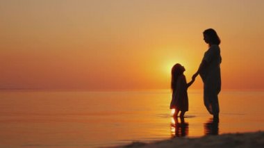Hamile bir kadın yavaşça onun en büyük kızı öper. Gün batımında deniz kenarında güzel siluetleri