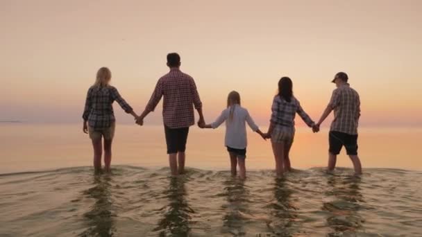 Un gruppo di amici con bambini che si divertono sull'acqua, tenendosi per mano camminando sull'acqua verso il sole al tramonto — Video Stock