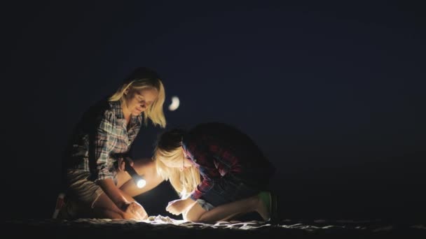 Mamma och dotter spelar tillsammans i sanden på natten. De lyser med en ficklampa som letar efter något. Äventyr och en lycklig tid grupp — Stockvideo