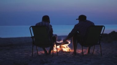İki arkadaş Marshmallow ateşi etrafında kızartıyorlar. Alacakaranlıkta nehir tarafından oturmak