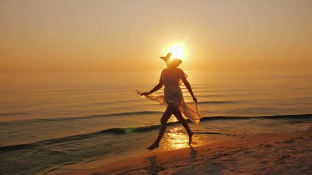 Μια γυναίκα σε ένα ελαφρύ φόρεμα απρόσεκτα περιπάτους κατά μήκος της ακτής στο ηλιοβασίλεμα — Αρχείο Βίντεο