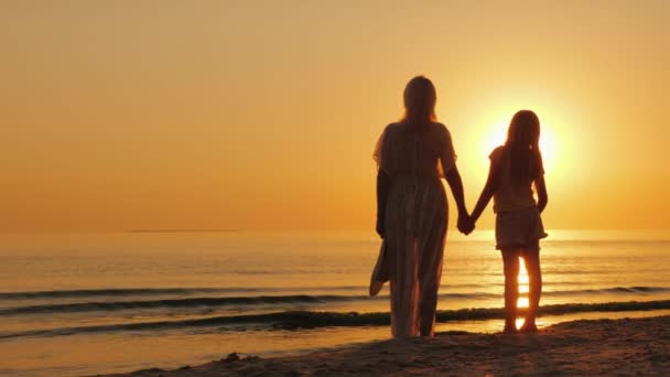 Una donna tiene per mano un bambino, sta insieme sulla riva del mare, guarda un bel tramonto. Madre con figlia — Video Stock