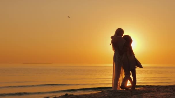 La mamma abbraccia dolcemente il bambino sullo sfondo del mare e del sole che tramonta. Silhouette di donna con bambino — Video Stock