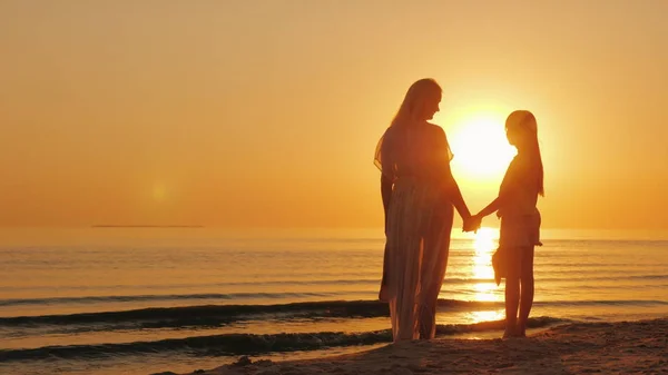 Máma a dcera se těší na krásný západ slunce nad mořem. Siluety ženy s dítětem poblíž vody — Stock fotografie