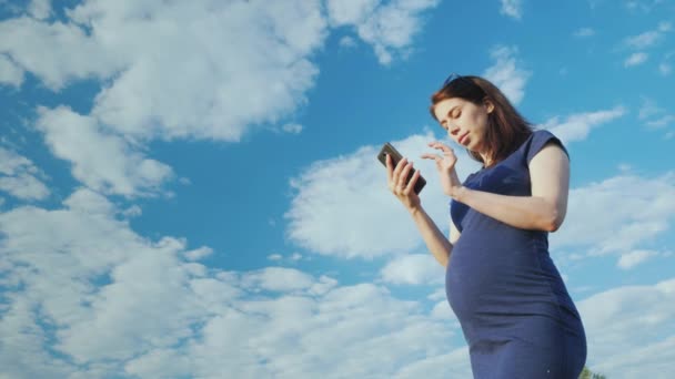 Una mujer embarazada utiliza un teléfono inteligente contra un cielo azul. Ángulo de visión inferior — Vídeo de stock