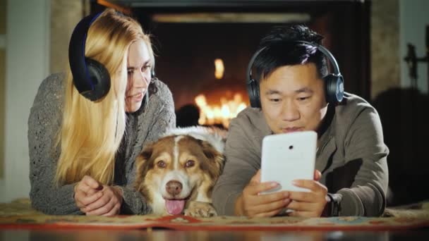 Um jovem casal está usando um tablet perto da lareira. Ao lado deles está o cão deles. Tecnologia e conforto no conceito de casa — Vídeo de Stock