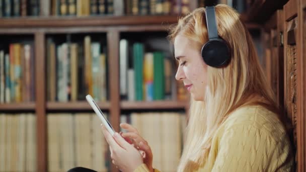 一个女学生正在图书馆里使用平板电脑。它是戴着耳机, 对书架的背景与书籍 — 图库视频影像