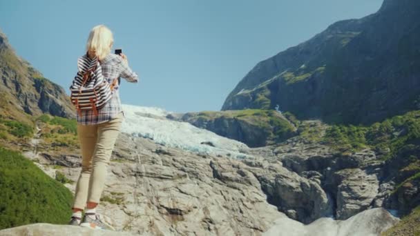 Uma mulher viajante ativa está em uma rocha, fotografa uma bela geleira na Noruega. Viagem pela Escandinávia — Vídeo de Stock