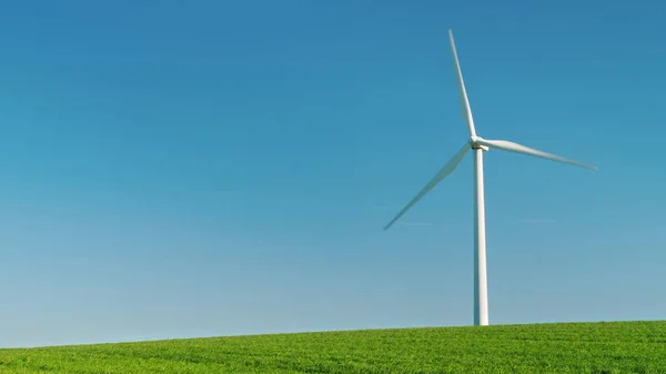 The industrial wind generator stands on a green hill. Against the background of the blue sky, an idylistic picture of pure energy