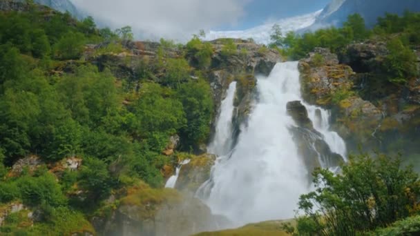 Hermosa cascada de las aguas del glaciar. Al fondo, la montaña con hielo en la parte superior es el glaciar Briksdal. La naturaleza de Noruega y Escandinavia — Vídeos de Stock