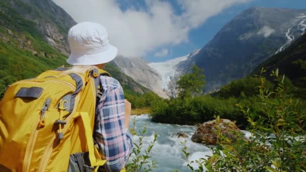 A tourist with a yellow backpack looks at a beautiful glacier at the top of the mountain. Briksdal glacier in Norway — Stock Video