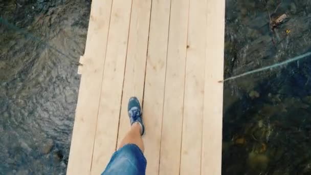 Een man loopt langs een smalle, wiebelende brug over de rivier, in het frame kan men alleen de benen — Stockvideo