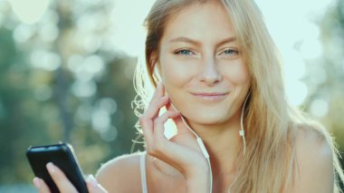 Genç bir kadın kulaklık müzik dinliyor, bir akıllı telefon kullanır. Güneş güzel saçlarını aydınlatır