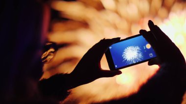 Bir kadın akıllı telefondan havai fişek atıyor. Işıklar gözlüklerine çok güzel yansıyor.