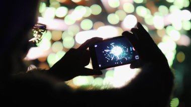 Bir kadın akıllı telefondan havai fişek atıyor. Işıklar gözlüklerine çok güzel yansıyor.